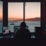 A person wearing a headset sits at a desk with multiple monitors, facing a sunset over the ocean through large window panes.