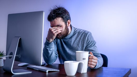 Bearded man in a gray sweater sits at a desk, head in his hand, looking stressed next to a computer and two coffee mugs.