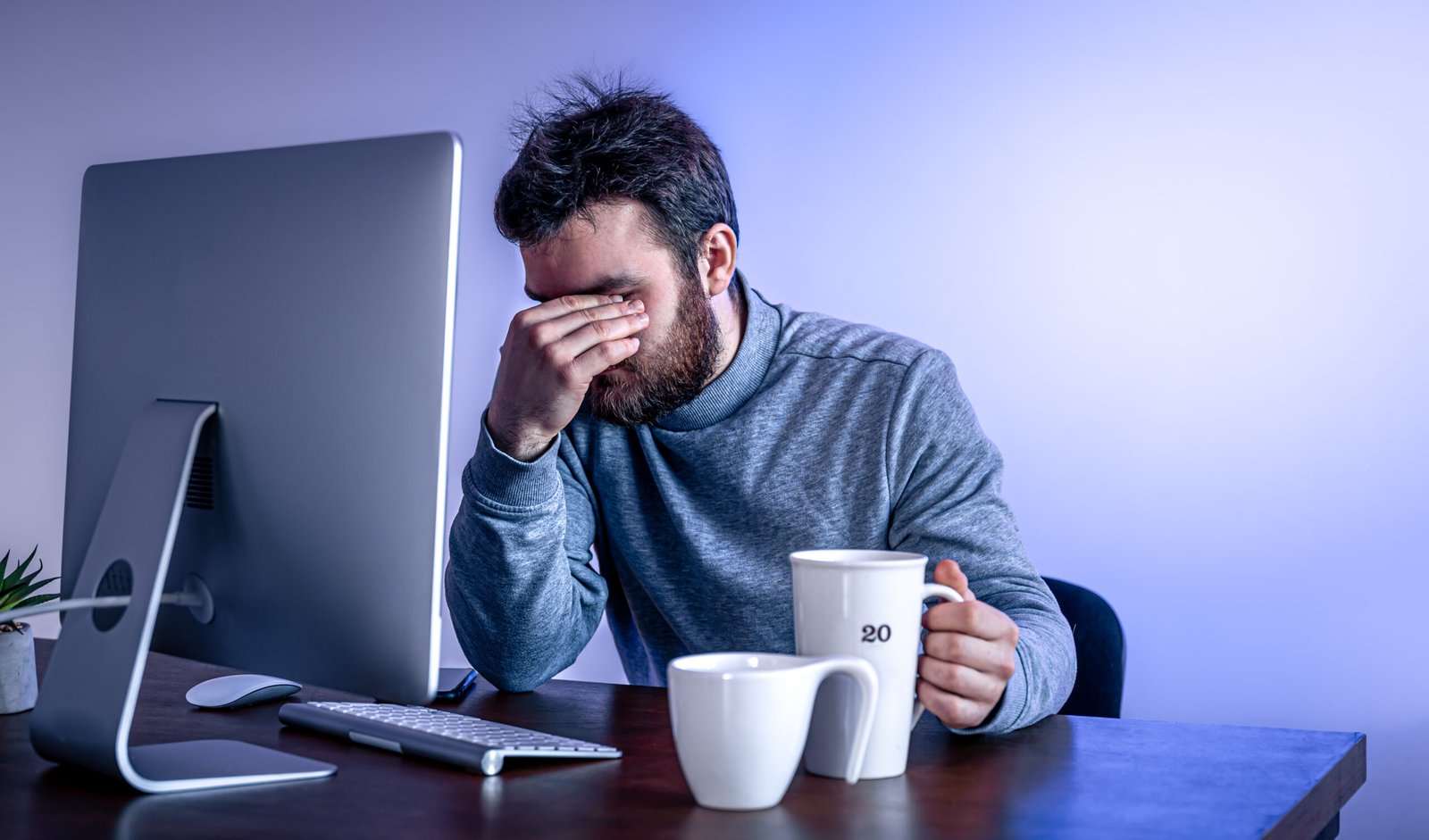 Bearded man in a gray sweater sits at a desk, head in his hand, looking stressed next to a computer and two coffee mugs.