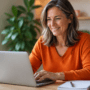 Smiling woman in an orange sweater typing on a laptop at a wooden desk in a bright home office
